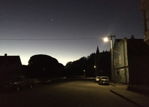 Night photo with streetlight at right, lighting the side of green wooden building and two cars underneath.  