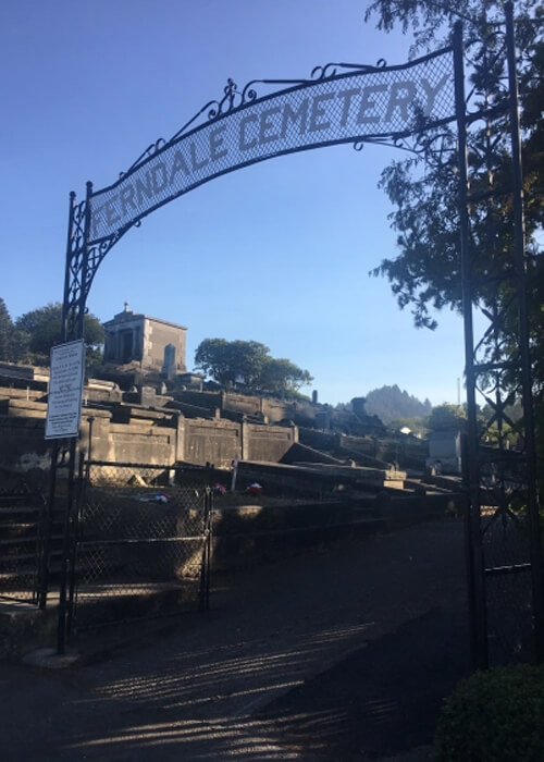 Cemetery gate with arch sign above reading "Ferndale Cemetery" and some tombs in the background.  Blue sky with no clouds in background.