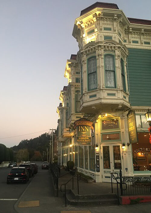 Corner shot at sundown of Victorian style two-story motel with sign hanging over door that says "Ferndale Inn."  Building painted green with white trim and ornate design trimmings.