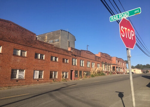 Block-long two story abandoned brick building with broken windows and stop sign at right with street sign that reads "Railroad Ave."  Blue sky in background.