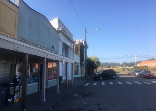 Row of old 1950's style single-story office buildings and a market at left, street disappearing into horizon at right.  Blue sky above with no clouds.