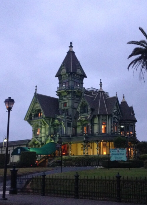 Large Victorian mansion at sundown with a series of towers and steeples, a few lights on inside.  Dark grey sky above.