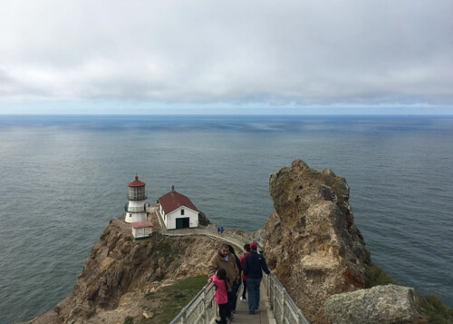 Outdoor stairway leading down rock ridge to a lighthouse, ocean behind the lighthouse and thick layer of clouds above in the sky.