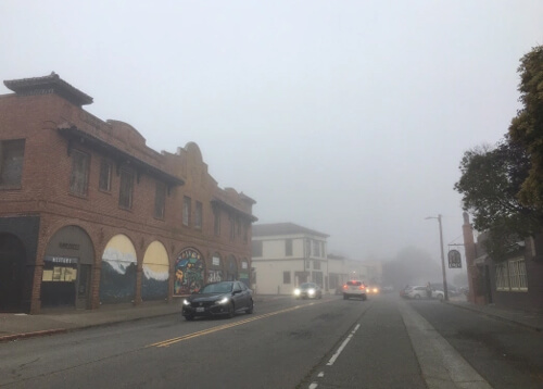 Fog covered street with large brick building on left side and a few car headlights peering through fog.