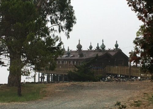 House on stilts over water with a series of round steeples on roof through a couple of trees.