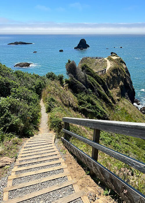 Stairs leading down a green covered cliffside to a lookout perch. Ocean beyond the perch with some rock islands. Blue sky above with a layer of clouds.