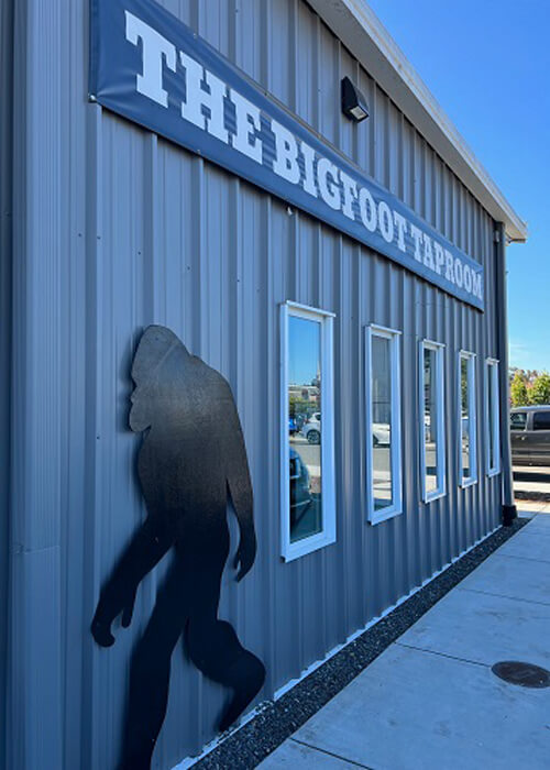 Side of grey aluminum building with sign reading "The Bigfoot Taproom" and a black-painted plywood Bigfoot figure underneath sign. Blue sky in background.