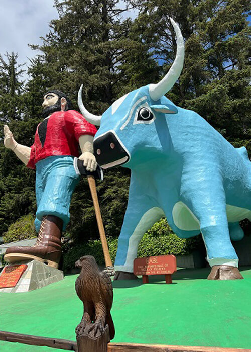 Looking up at giant Pal Bunyan statue at left and his blue ox at right, some trees and blue sky behind them.