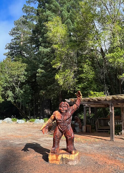 Wooden Bigfoot statue with one arm raised in air, some trees in background. Blue sky above with a few clouds.