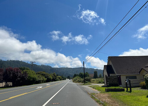 Road at left eventually disappearing into forested hills, small church at right. Blue sky with some clouds.