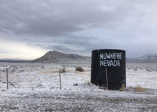 Snow covered desert terrain and hills in background, rusty cylinder tank at right with sign painted on reading "nowhere Nevada." Grey cloudy sky above.