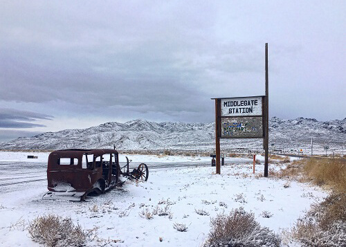 Remains of rusty early-1920's coupe buried in the snow in the ground at left, sign at right reading "Middlegate Station." Snow covered mountains in background and grey sky.