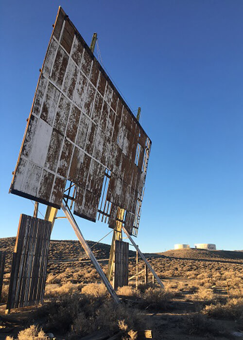 Abandoned drive-in movie theatre screen in the desert, with part of the screen missing. Blue sky above.