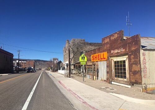 Abandoned Shell gas station at right, with rusty exterior and yellow and orange sign saying "Shell." Road at left disappears into desert mountains. Blue skies above.