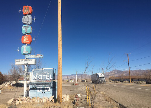 Old motel sign at left with vertical stacked multi-colored letters reading "motel." Street at right with large white truck driving by and blue sky above.