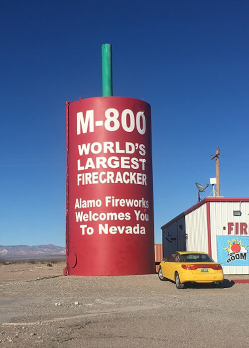 Five-story high red cylinder with white lettering that reads "M-800, World's largest Firecracker," with desert terrain in background and yellow car parked in front. Blue sky with no clouds.