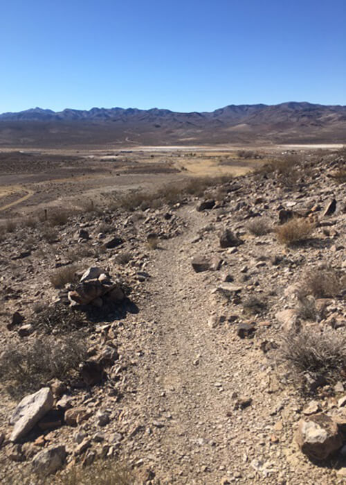 Trail pointing ahead in between desert rocks, desert brush and tumbleweeds. Mountain ridge in background and blue sky above.