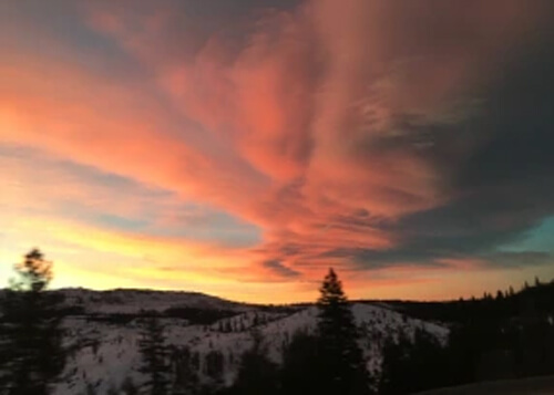 Orange clouds at sunrise over a snow covered mountain ridge and forest trees.