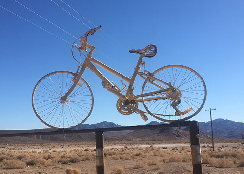 Ornamental bike pained white and perched on top of gate, desert scenery and a mountain ridge in background, blue sky above.