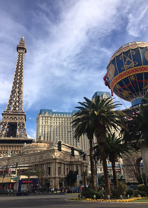 Replica of Eiffel Tower at left, and replica of hot air carnival balloon at right, with palm trees and a thirty-story hotel in between. Blue sky with scattered clouds.