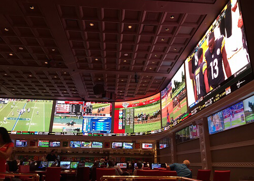 Inside a sports betting casino, with row of ten-foot TV screens wrapped around inside of building. Some people below in chairs watching games.