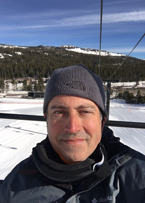 Picture of Mark Loftin's face on ski lift, wearing ski cap, snow and forest covered mountains in background.