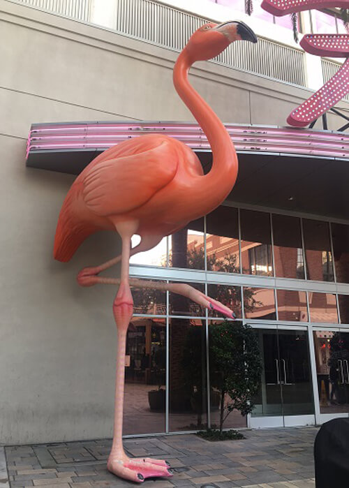 Giant two-story flamingo perched on one leg outside The Flamingo Hotel. Hotel entry at right.