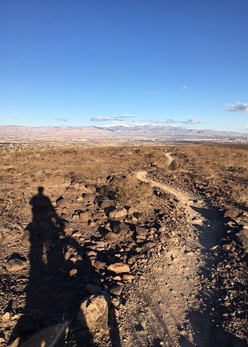 Shadow of me on mountain bike overlooking desert trail ahead with sparse desert terrain of dirt and rocks in early morning light. Blue sky with a few clouds above.