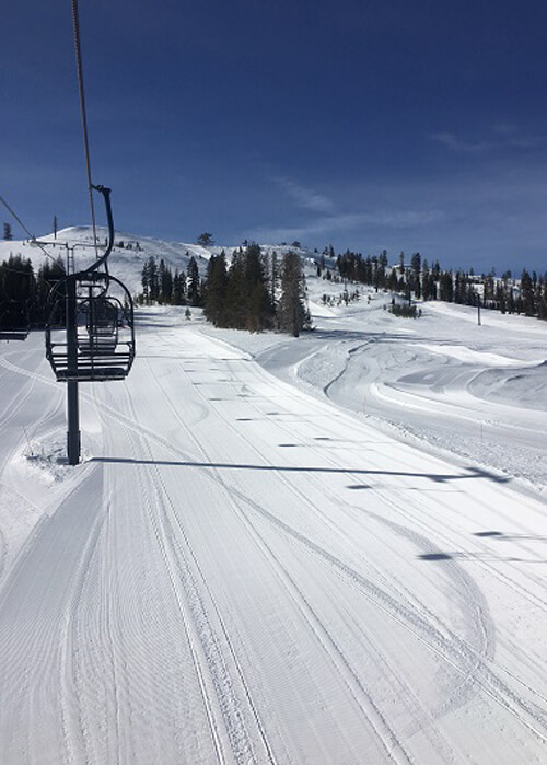 View from ski lift showing groomed snow underneath the chair lifts ahead. Some pine trees in background. Clear blue sky.