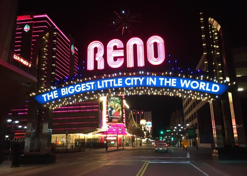 Night photo of famous Reno sign, saying "Reno" in neon pink and "biggest little city in the world beneath" in an arch lighting structure over the street, casino lights in the background. 