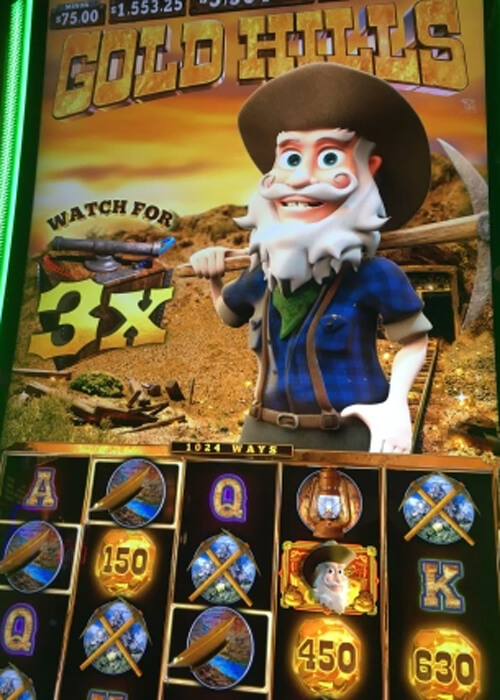 Close up photo of slot machine, reading "Gold Hills" with a bearded gold miner smiling and slot reels below in various gold mining tools and gold coins.