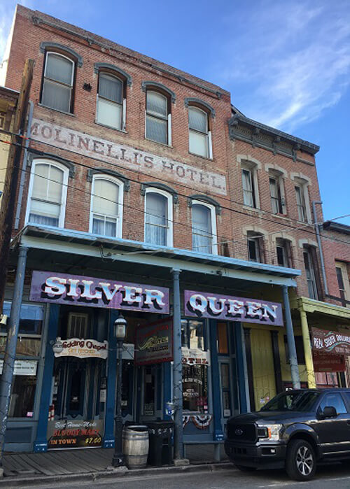 Three-story brick hotel looking from the gold rush era, with sign above first floor saying "Silver Queen." Blue sky above.