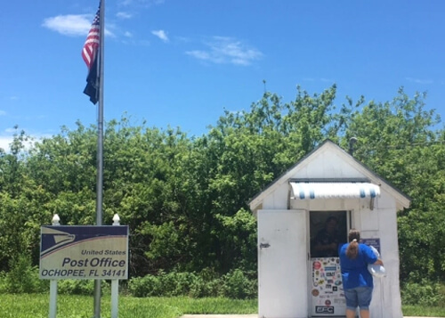 alt="Small white shack at right with lady in front, post office sign at left, with green trees behind. Blue sky above."