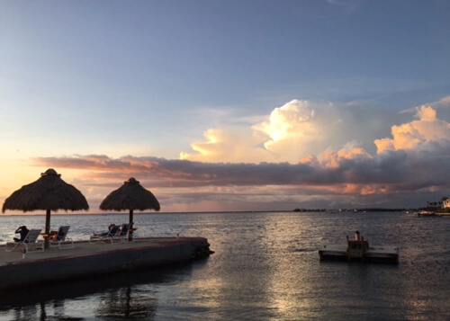 alt="Two umbrella covered tables at left, overlooking calm water reflecting the bright white and orange clouds above at sundown. A few people in beach chairs below the huts."