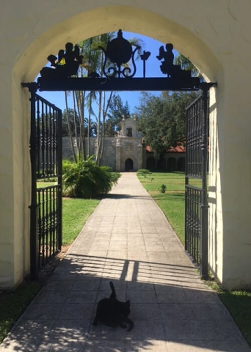 Arched doorway lined with black iron design at top and black iron gates. Through door is stone monastery building in the distance.