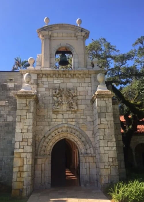 White and grey stone monastery corner section, with bell tower and arched doorway. Blue sky behind building.