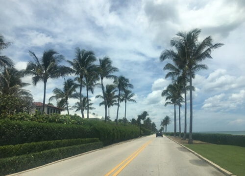 Road lined with palm trees and the edge of a stucco roof home on left and ocean on right. Cloudy blue skies behind the road.