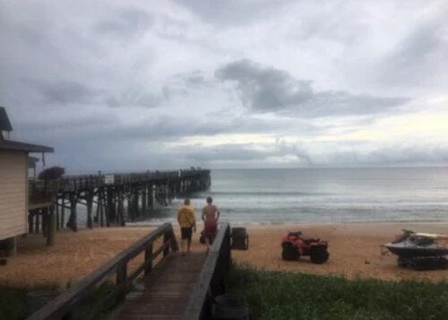 Wooden walkway with two people standing at end overlooking beach and ocean, and boat at bottom right. Grey clouds in the sky above.