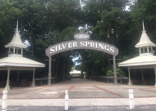 Arched sign entryway reading Silver Springs flanked by two white canopies for shade. Thick green trees behind the sign, and pavement below.