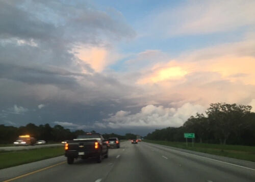 Driving on freeway with green grass and trees on both sides, cars in front and a sundown sky with layers of white and dark clouds