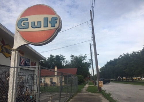 Orange Gulf gas station sign at upper left, with road at right and grey sky above. Dark green trees line the street.