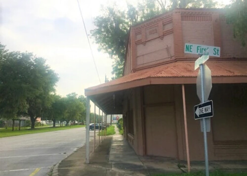 Street corner with stop sign and sign reading "NE First St," orange painted single story building at corner.