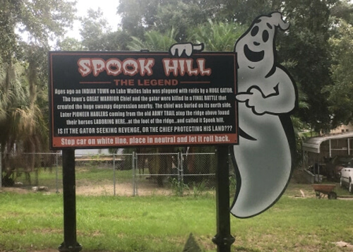 Sign displaying the history of Spook Hill, with a painted ghost pointing to the sign. Green grass below the sign, and some thick green trees behind it.