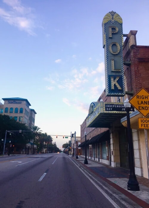 Vertical blue sign at upper right saying "Polk,' with shop lined street disappearing into horizon straight ahead at sunrise. Light blue sky above with a few clouds.