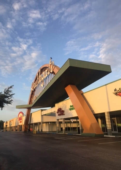 Large multi-story orange arch in front of a shopping mall, blue sky with scattered clouds above the sign.