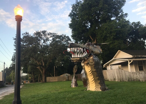 Dragon head protruding from a front lawn with big white teeth in center of picture. Some green trees behind dragon, and street lamp at left. Blue sky with scattered clouds above.