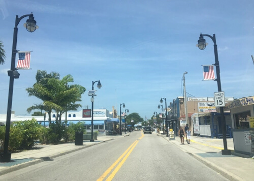 Street leading into a town with street lamps on both sides and American flags hanging from them. Some shops and trees on both sides of street. Blue sky above.