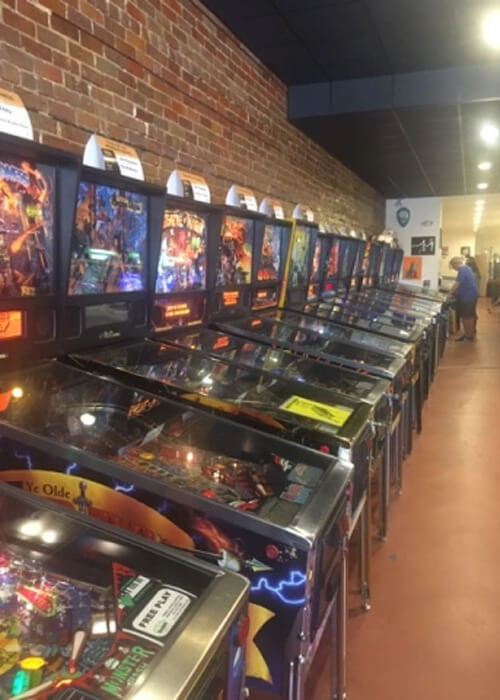 A row of pinball machines on left side and brick wall behind them. Walkway at right with a few pinball players in the distance.