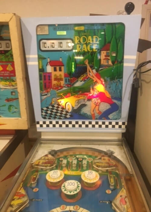 Close up of pinball machine from 1969 that says "Road Race" at top with a couple women clapping and cheering on a two seater sports car painted on the machine.