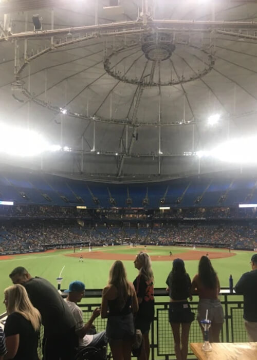 View of inside of indoor baseball field from behind center field, with baseball field below and some spectators watching from edge of the fence. Grey dome tower above with bright white lights hanging from it.